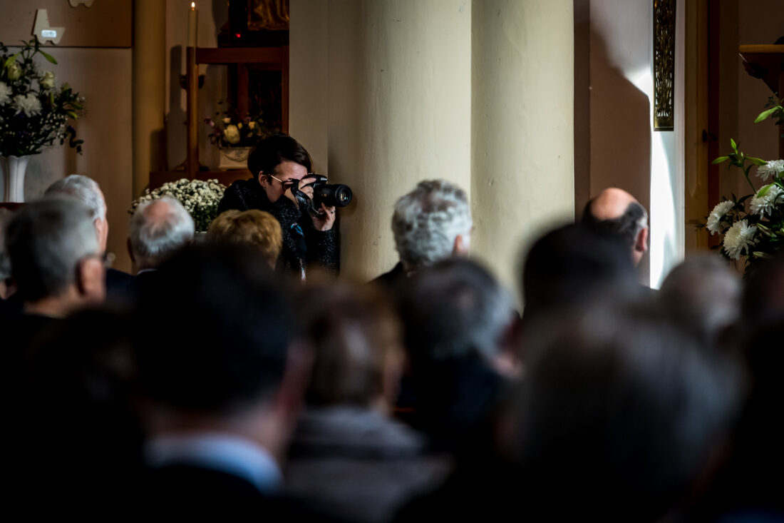 Uitvaartfotograaf werkt onopvallend langs een pilaar tijdens een kerkdienst